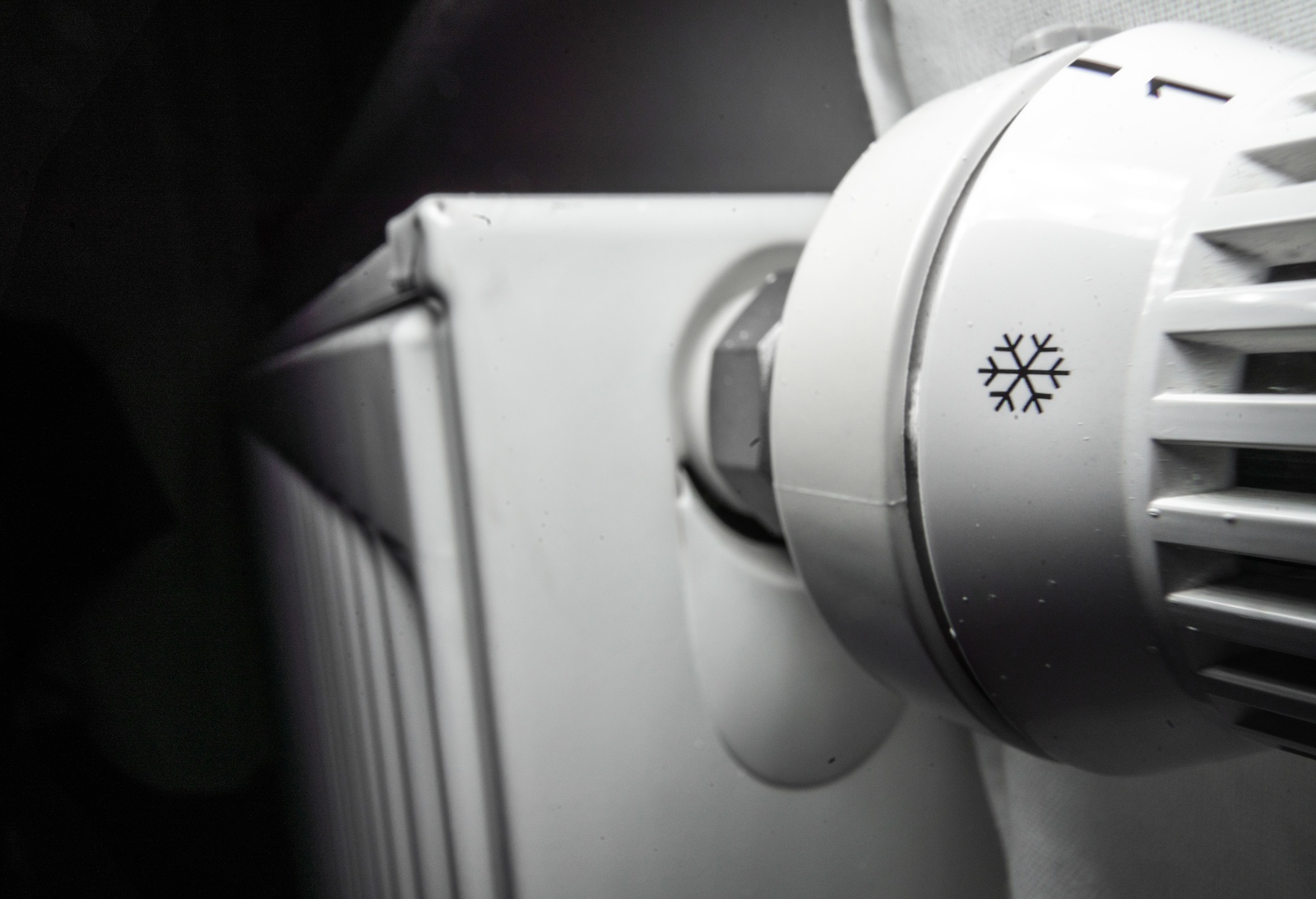 A closeup of the white thermostat knob of radiator, with a snowflake symbol visible on the knob, and the radiator cloaked in shadow