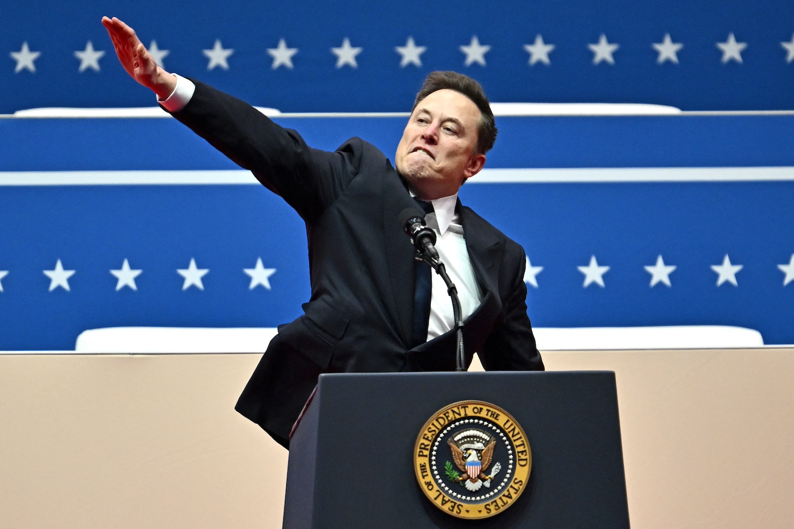 Elon Musk stands at a podium bearing the seal of the president of the United States with his right arm raised above shoulder height and his wrist and palm extended