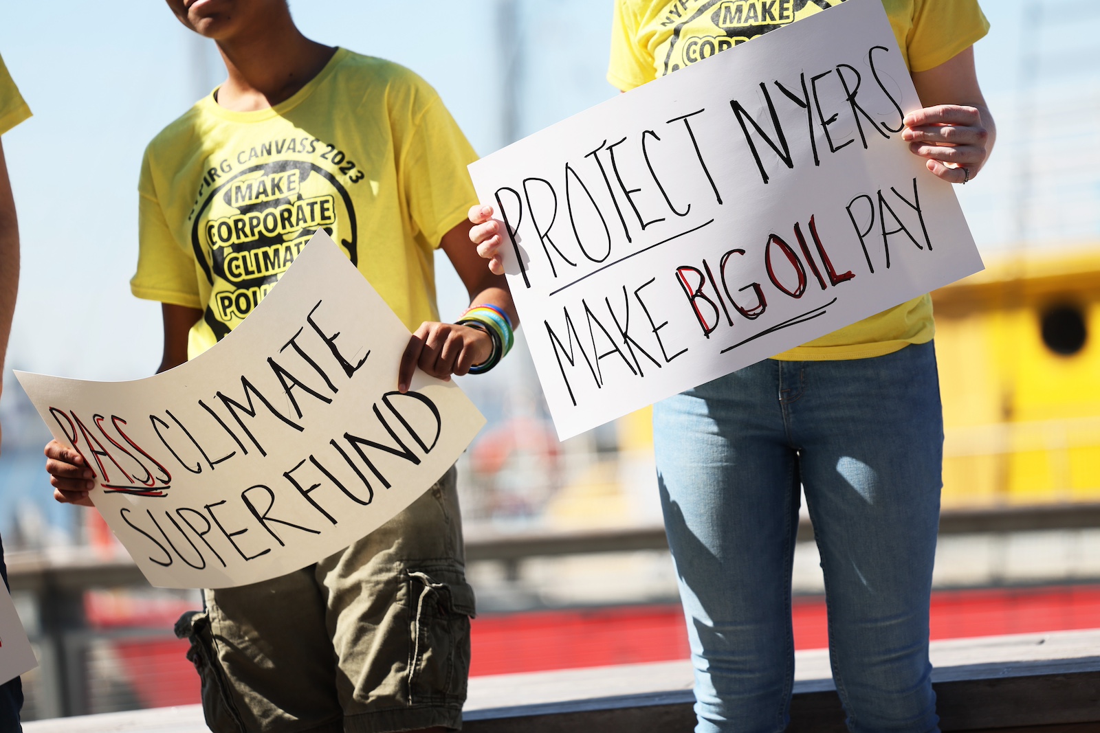 Two people, viewed from the neck down and wearing yellow t-shirts, hold signs saying 'Pass Climate Superfund' and 'Protect NYers Make Big Oil Pay'